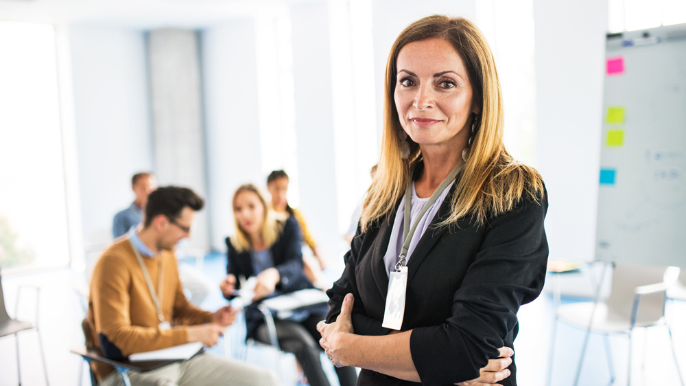 a female coach standing in a board room, arms crossed. A group of businesspeople sitting, making notes