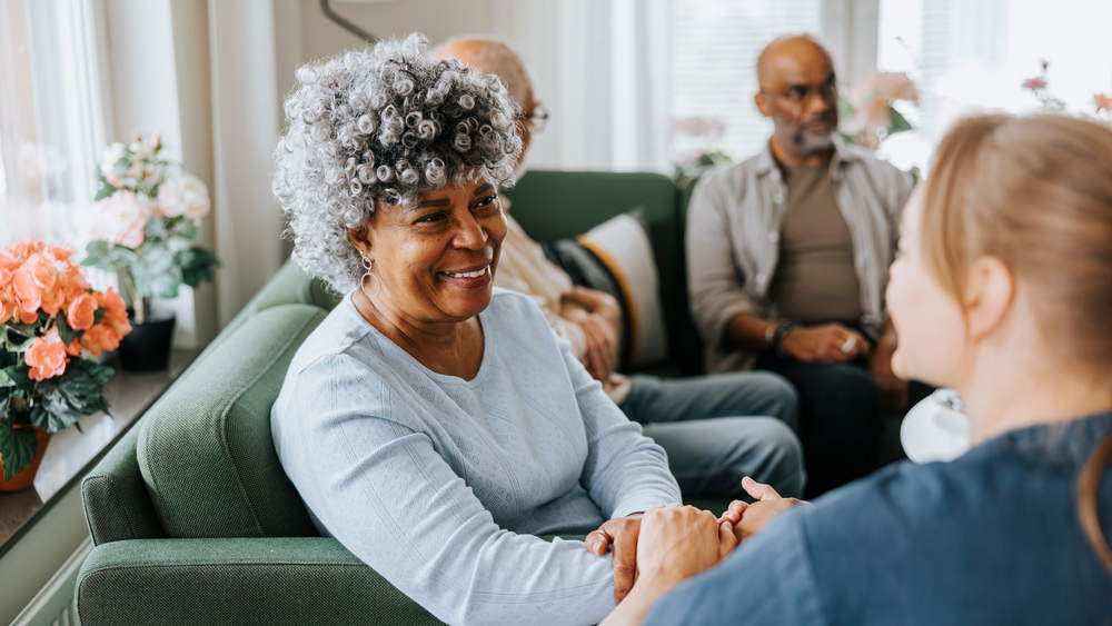 miling senior woman sitting on sofa and talking with healthcare expert at nursing home
